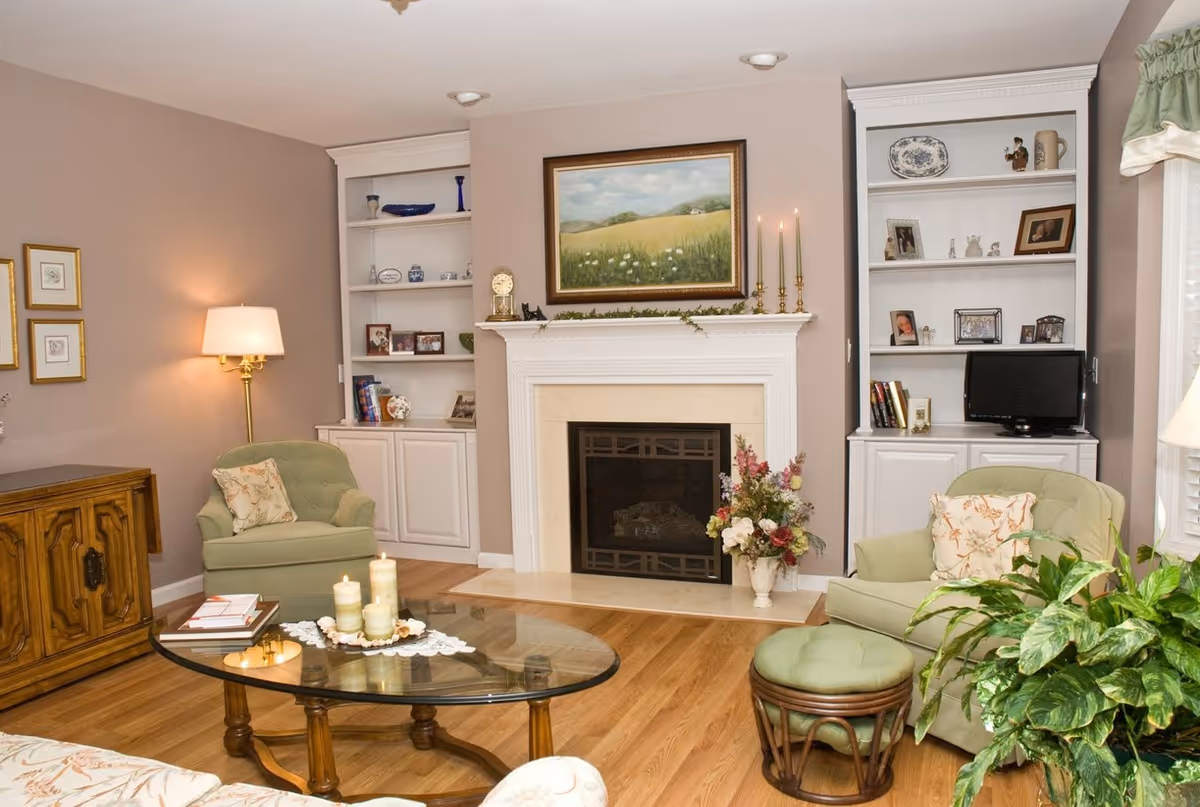 A cozy living room with two green armchairs, a glass coffee table with candles, a wooden cabinet, built-in white shelves with decorative items, a fireplace with a painting above it, and a large green plant near a window with white blinds and green curtains.