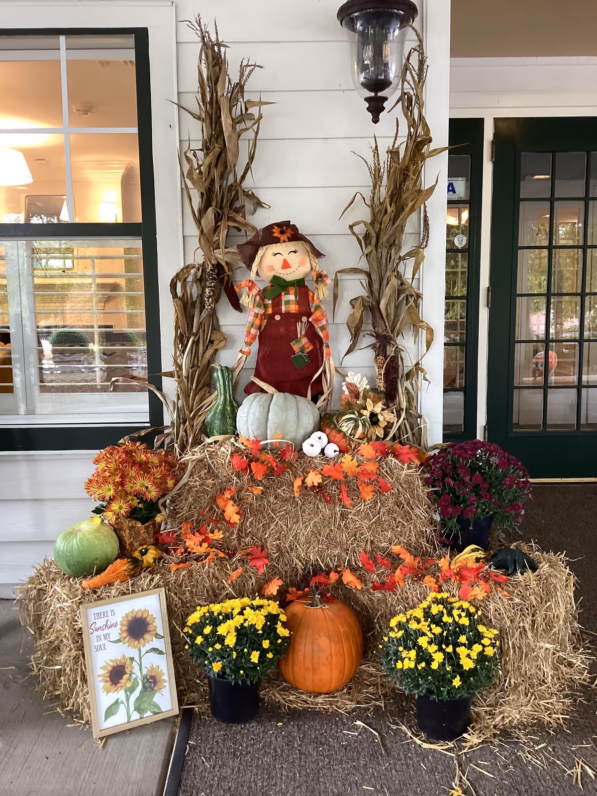Fall-themed entrance display with a scarecrow, hay bales, pumpkins and potted mums set against a building doorway.