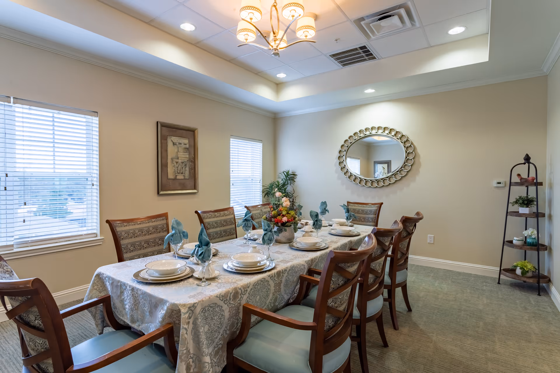 A well-lit dining room with a long rectangular table covered with a patterned tablecloth. The table is set with plates, bowls, glasses with folded blue napkins, and a floral centerpiece. There are eight wooden chairs with cushioned seats around the table. The room has two windows with blinds, a decorative framed picture on one wall, an oval mirror with a circular pattern frame on another wall, and a corner shelf with small plants and decorative items. The ceiling has recessed lighting and a chandelier.