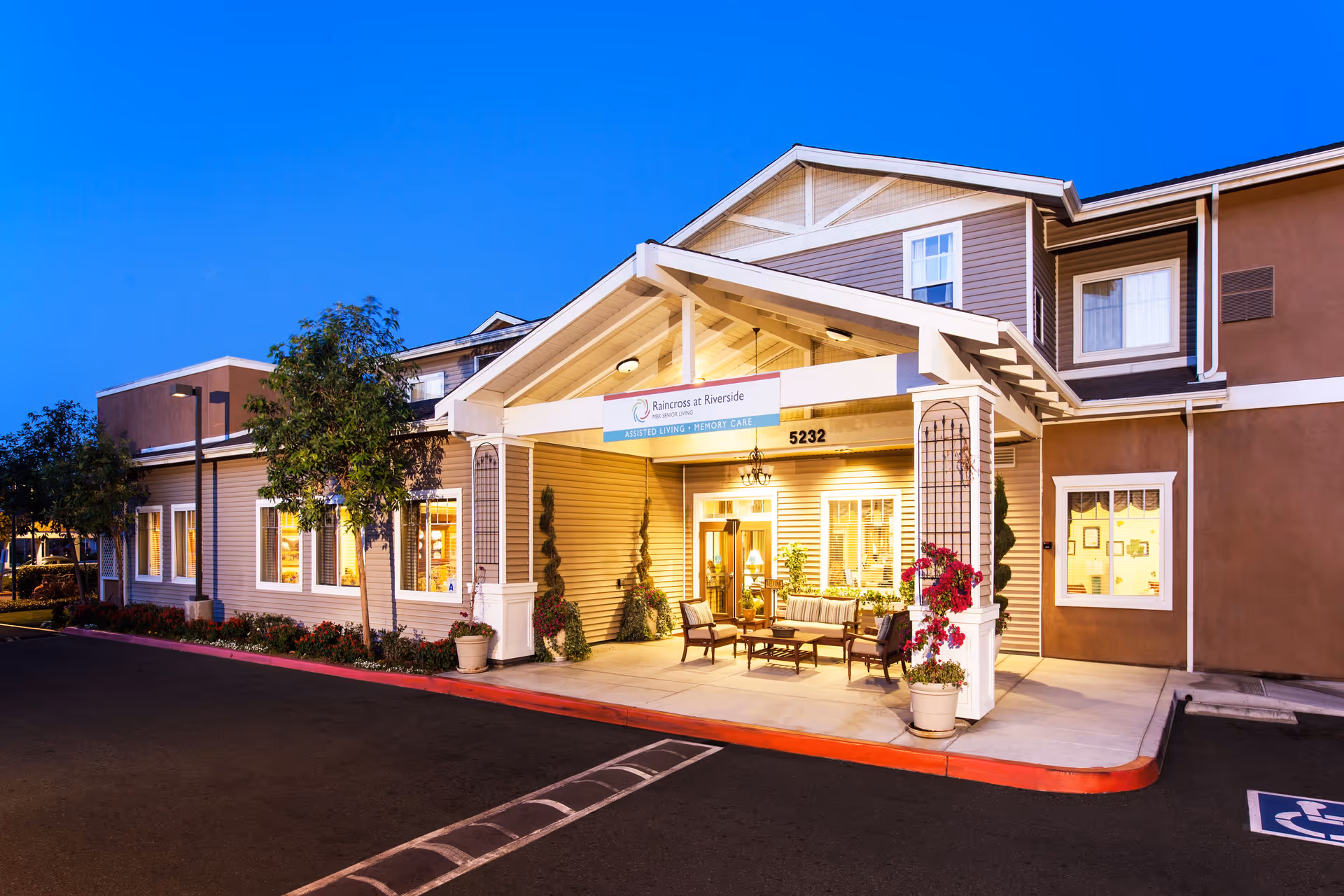 Exterior view of a senior living facility named Raincross at Riverside during twilight. The building has a covered entrance with seating and potted plants, illuminated warmly from inside. The address number 5232 is visible above the entrance.