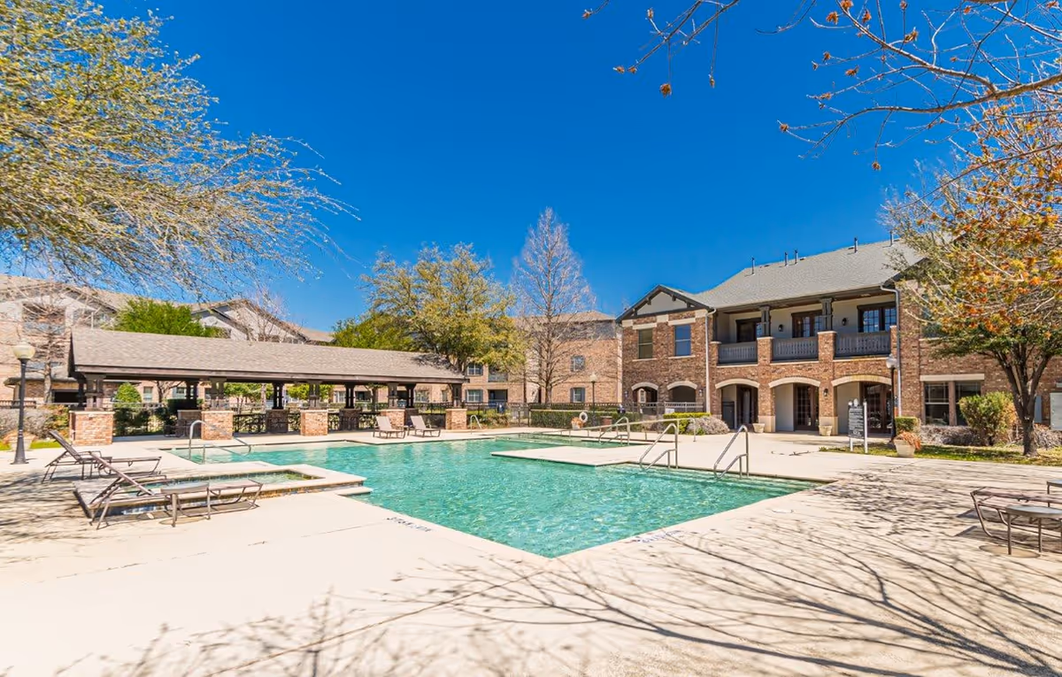 Outdoor swimming pool with lounge chairs in a courtyard in front of a two-story brick apartment building under a clear blue sky.