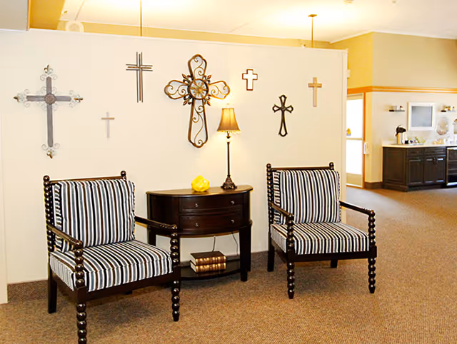 A cozy seating area with two wooden chairs featuring black and white striped cushions. Between the chairs is a small dark wooden table with a lamp and decorative items. The wall behind the chairs is decorated with various styles of crosses. In the background, there is a kitchenette area with cabinets and a coffee station.
