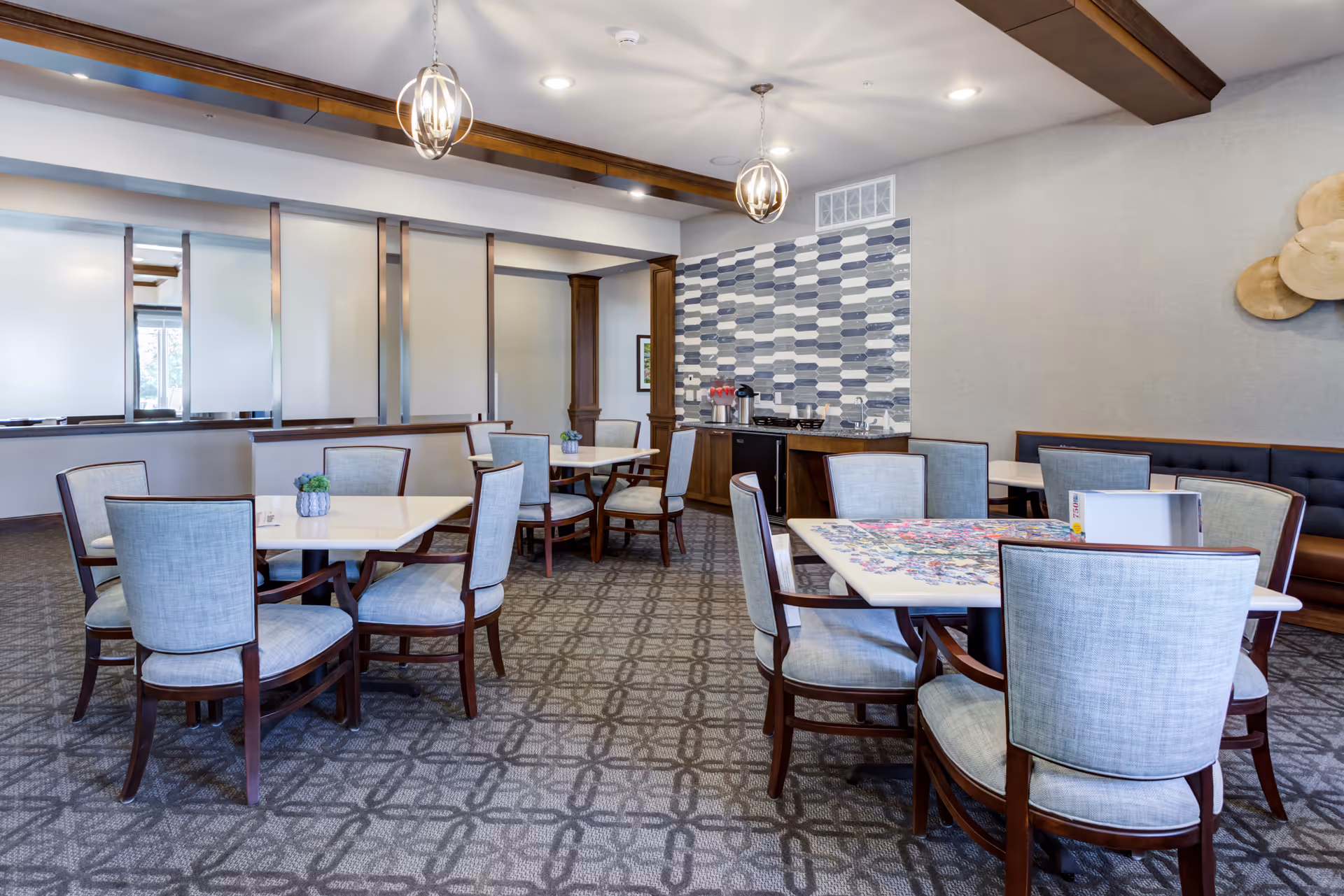 A bright and clean dining room with multiple square tables and cushioned chairs arranged neatly. One table has a partially completed colorful puzzle on it. The room features patterned carpet, modern pendant lights, a decorative tiled wall, and a small counter area with coffee dispensers and cups.