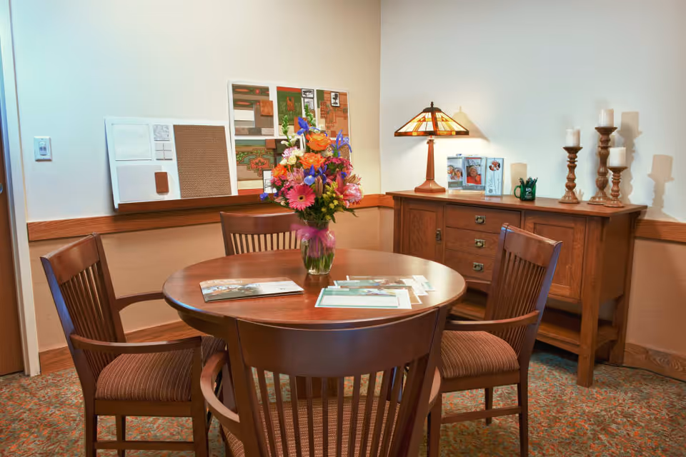A cozy interior room with a round wooden table surrounded by four wooden chairs with cushioned seats. On the table is a vase with a colorful bouquet of flowers and some brochures or papers. Against the wall is a wooden sideboard with a decorative lamp, framed photos, a green mug with pens, and several candle holders with white candles. The walls are light-colored with a wooden chair rail and carpeted floor with a patterned design.
