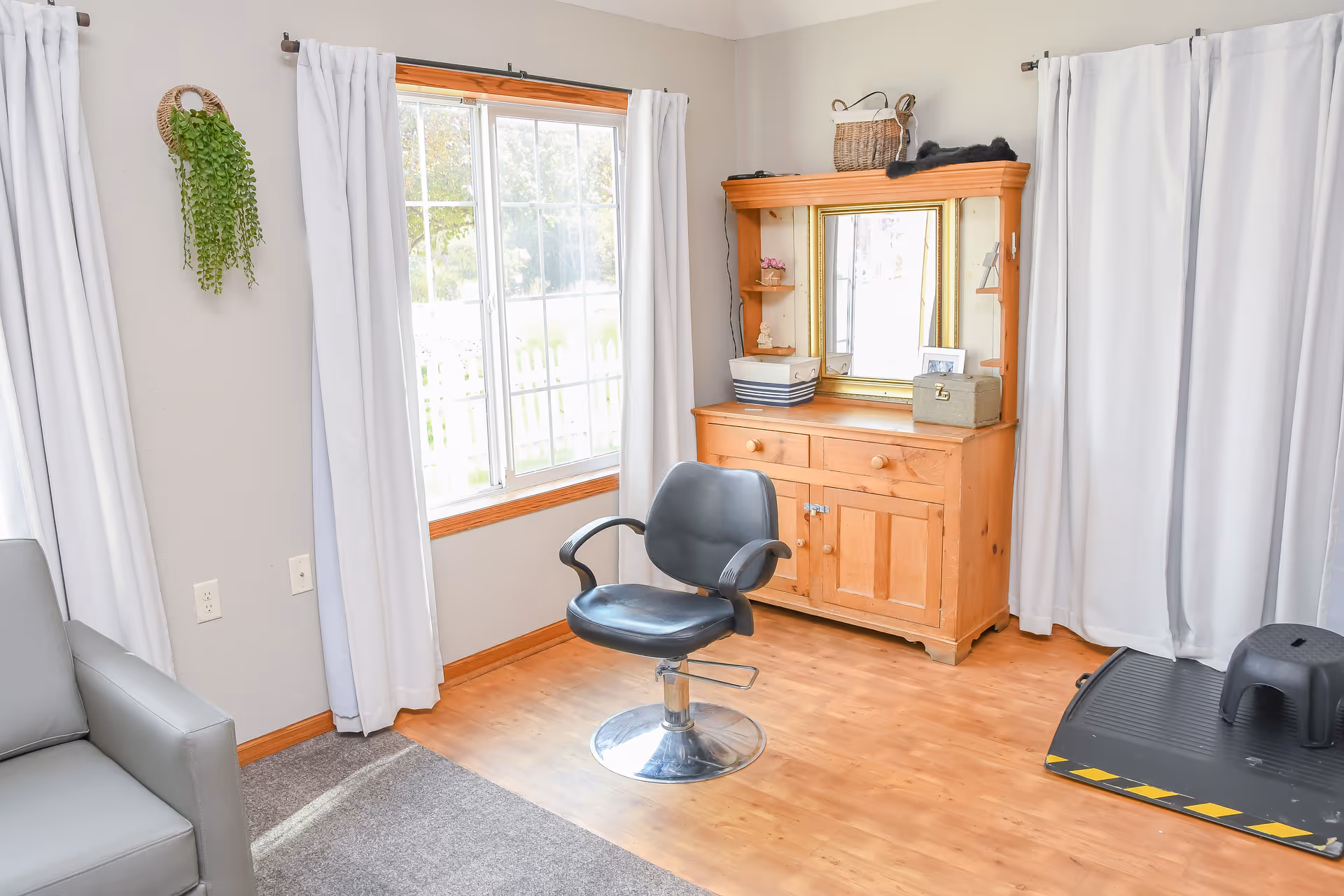 Bright room with a salon-style chair in front of a wooden dresser with mirror, a large window with white curtains, and light wood flooring.