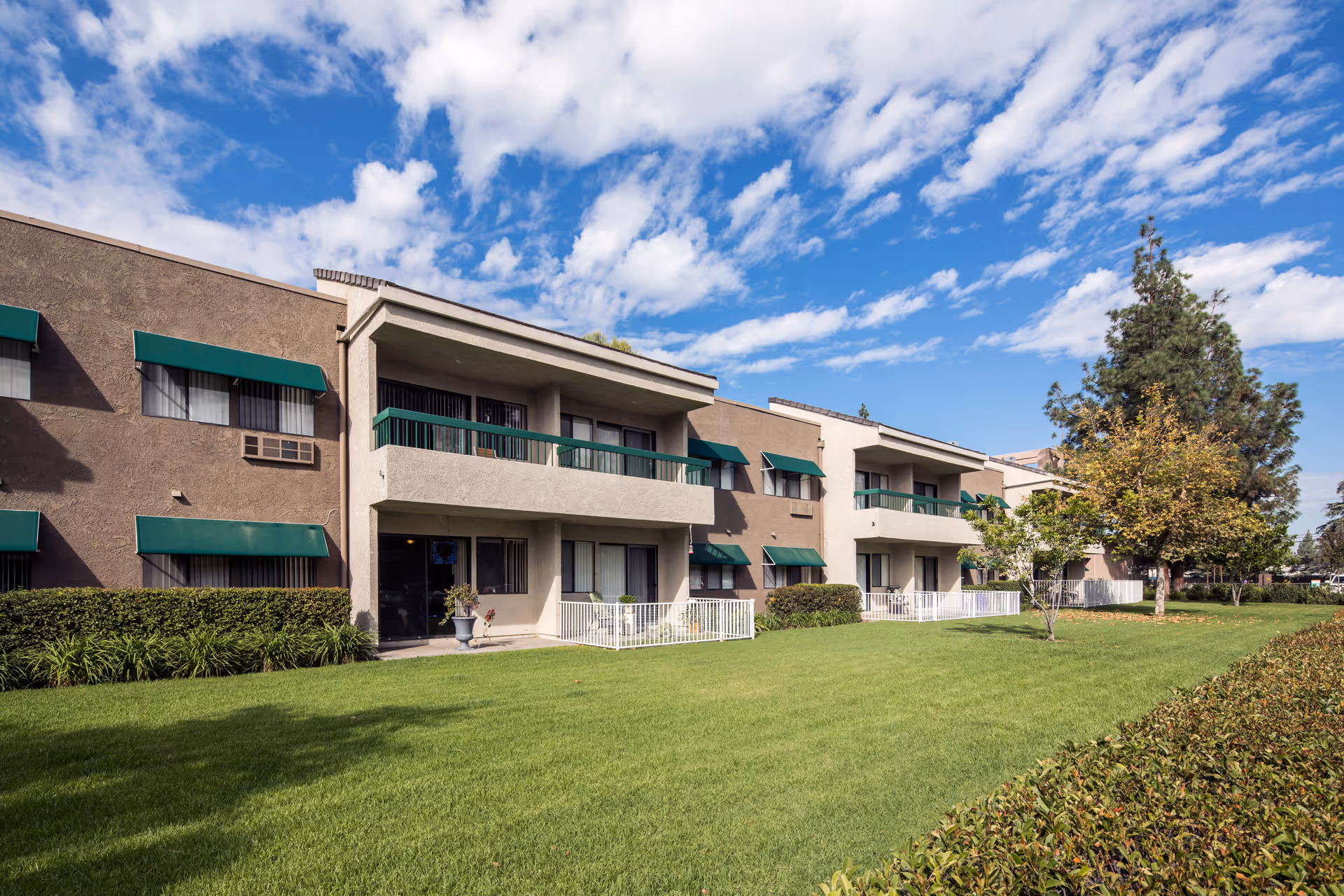 Exterior view of a two-story residential building with balconies and green awnings over the windows. The building is surrounded by a well-maintained lawn, bushes, and trees under a partly cloudy blue sky.