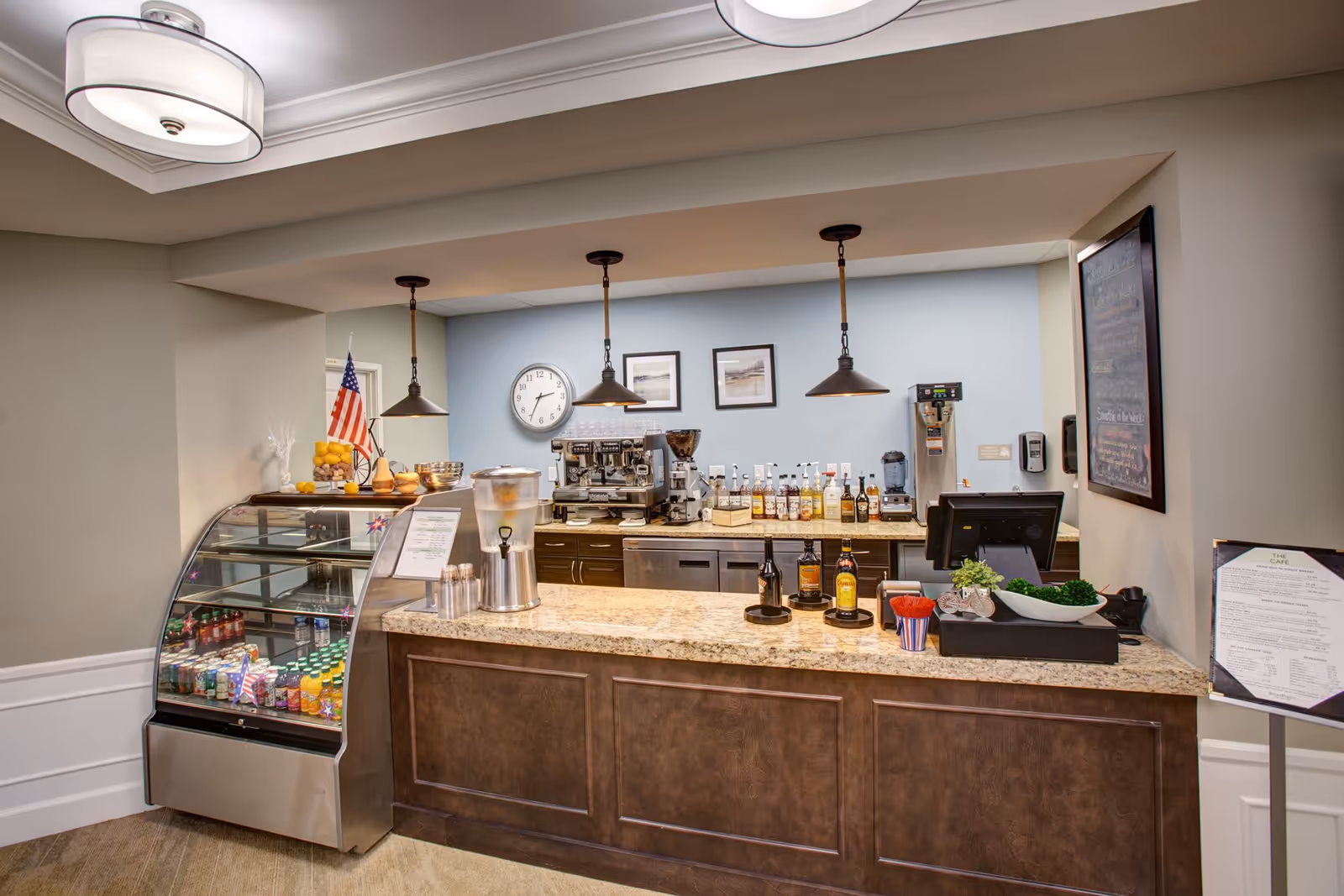 A well-lit coffee bar inside a senior living facility with a display case of drinks, a granite counter, espresso machine, and pendant lights.