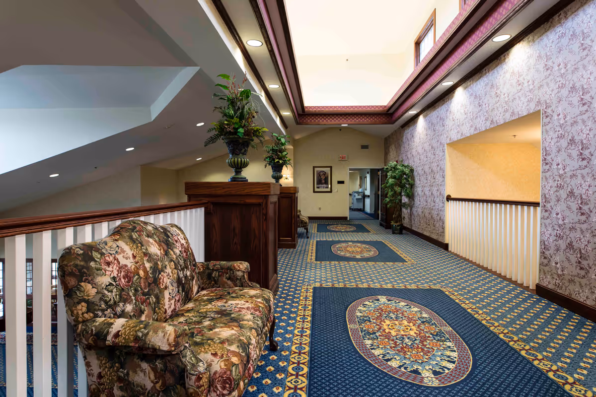 An upstairs interior corridor with patterned carpeting, a floral upholstered loveseat, decorative plants, and a skylit ceiling.
