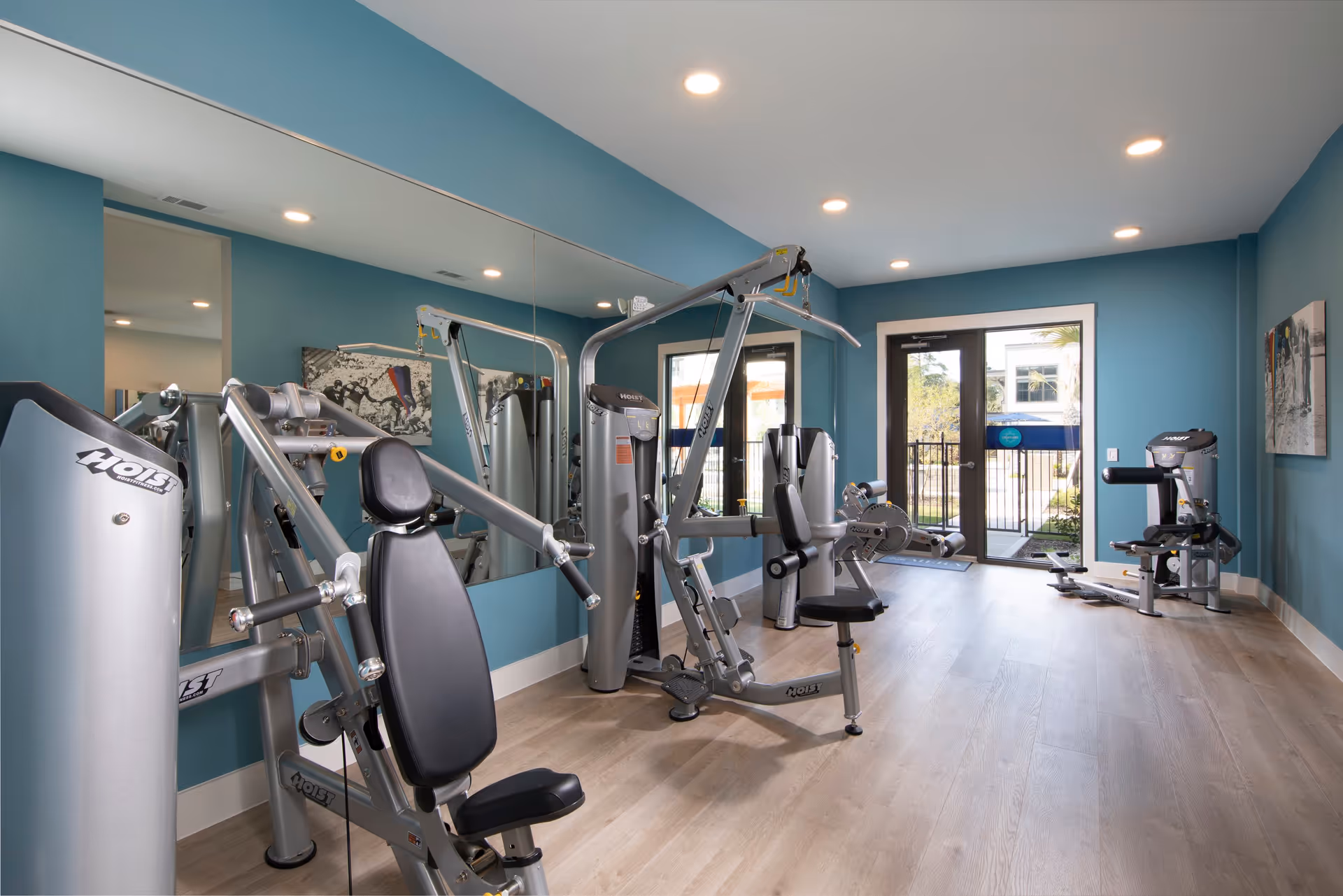 A small fitness room with blue walls and light wood flooring. The room contains several pieces of Hoist brand exercise equipment, including weight machines and a rowing machine. A large mirror covers one wall, reflecting the equipment. There is a glass door at the far end leading outside, allowing natural light to enter. The room is well-lit with recessed ceiling lights.