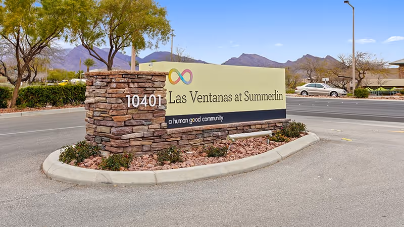 Stone and brick sign at the entrance of Las Ventanas at Summerlin community, with the address 10401 and the tagline 'a human good community'. The sign is surrounded by landscaping with small plants and rocks, with a road and mountains visible in the background under a clear sky.