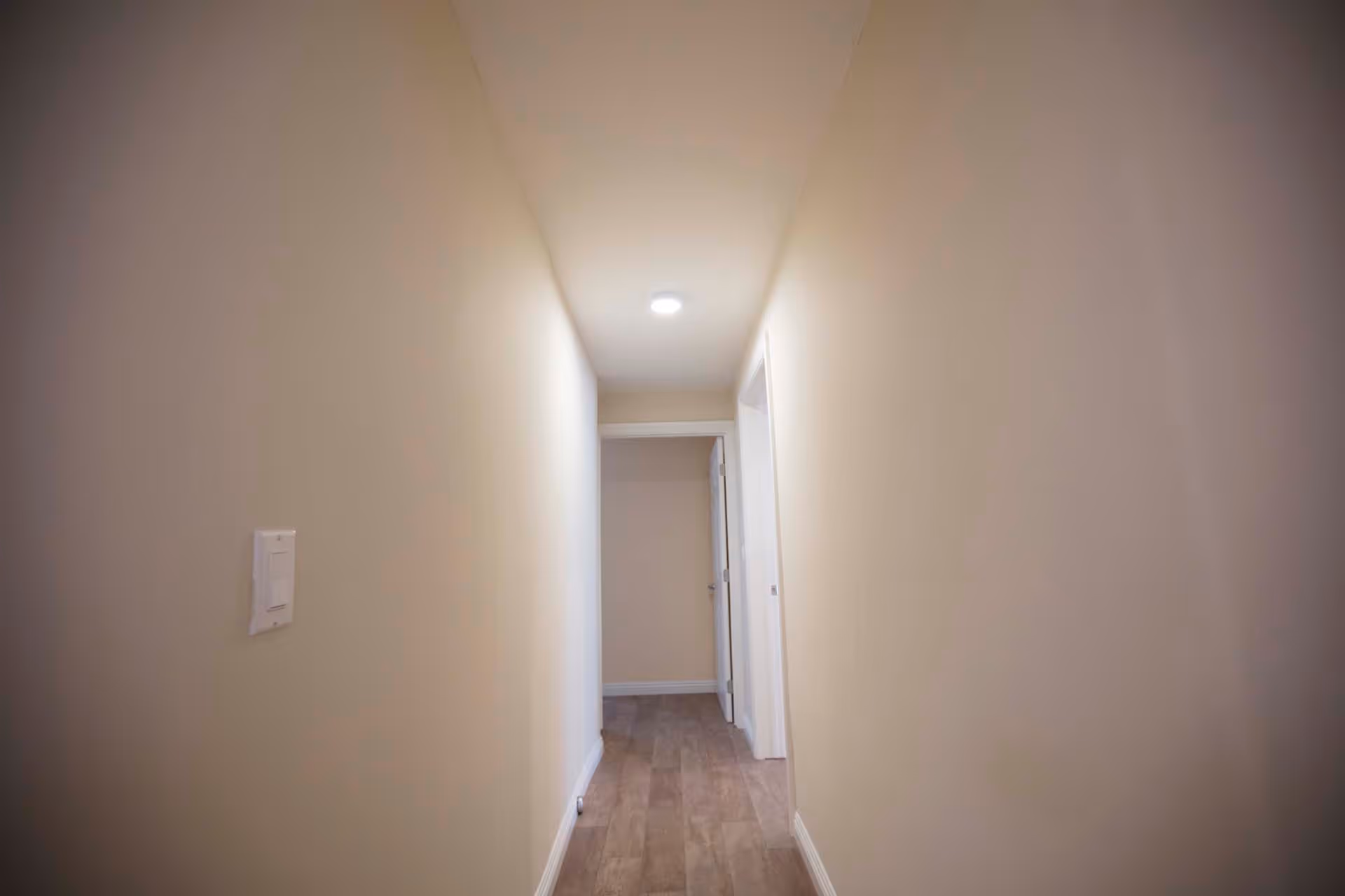 A narrow, well-lit hallway with beige walls and wooden flooring. There is a light switch on the left wall and a ceiling light fixture illuminating the corridor. At the end of the hallway, there is an open doorway leading to another room.