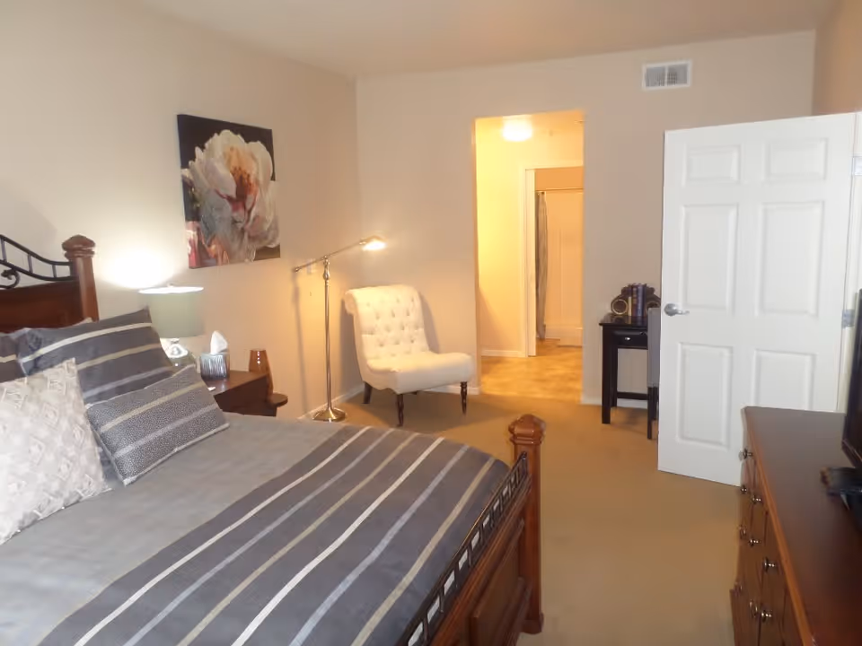 A bedroom with a wooden bed frame and striped bedding, a nightstand with a lamp and tissue box, a white upholstered chair with a floor lamp beside it, a dresser with a TV on top, and an open door leading to a bathroom.