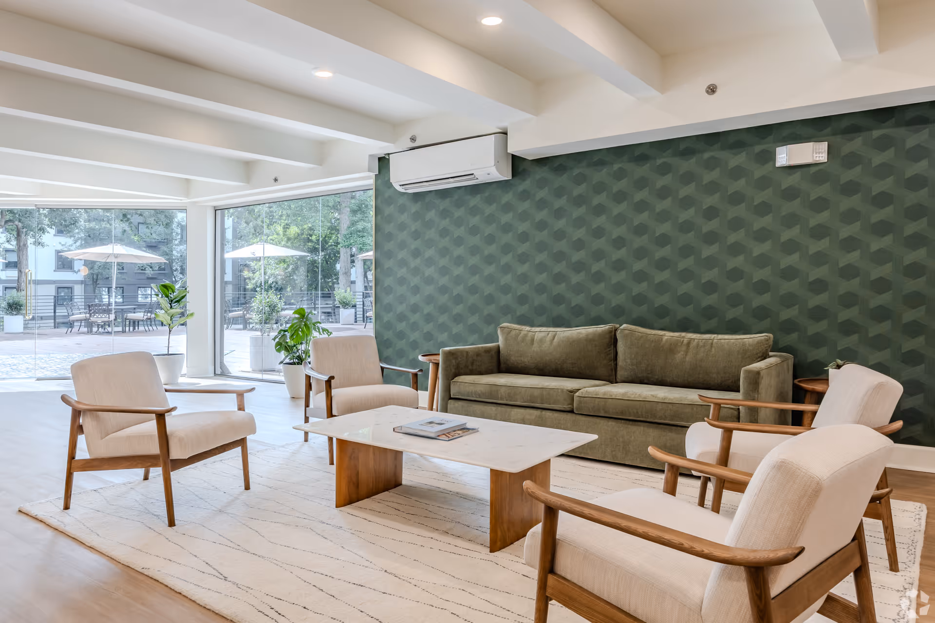 A modern living room with a green patterned accent wall, a green sofa, four beige armchairs with wooden frames, and a rectangular white coffee table on a light-colored rug. Large windows provide a view of an outdoor patio with tables, chairs, and umbrellas.