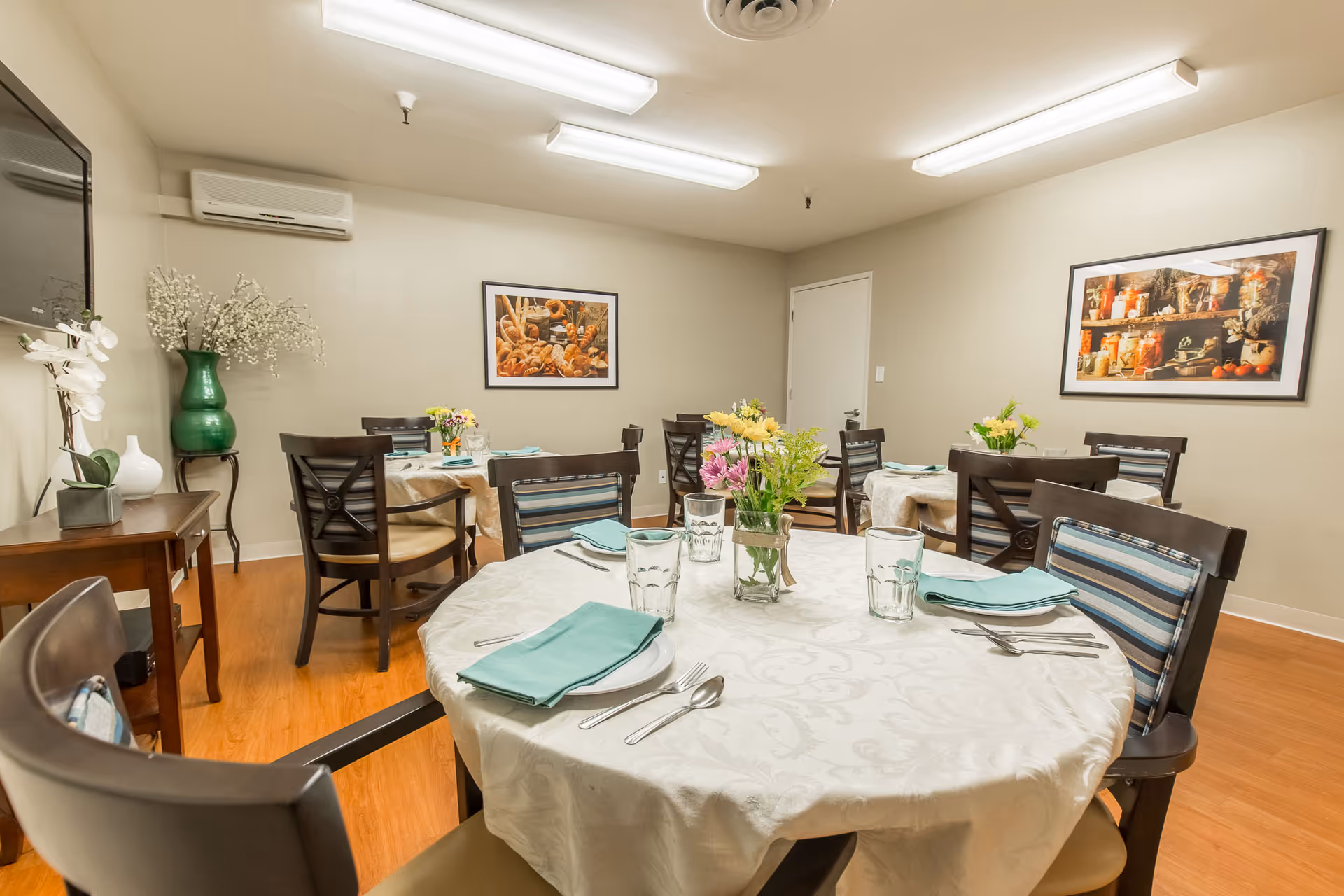 A dining room with round tables covered in white tablecloths, each set with plates, glasses, silverware, and teal napkins. There are dark wooden chairs with striped cushions around the tables. The room has light-colored walls with framed pictures of food, a wooden side table with decorative vases and flowers, and bright overhead fluorescent lighting.