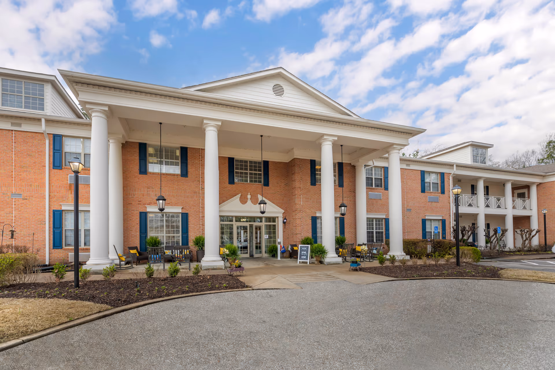 Front exterior view of a two-story brick building with large white columns supporting a covered entrance. The building has blue window shutters and several benches with yellow cushions near the entrance. There are lamp posts along the driveway and a sign near the entrance that reads 'Welcome Home'. The sky is partly cloudy.