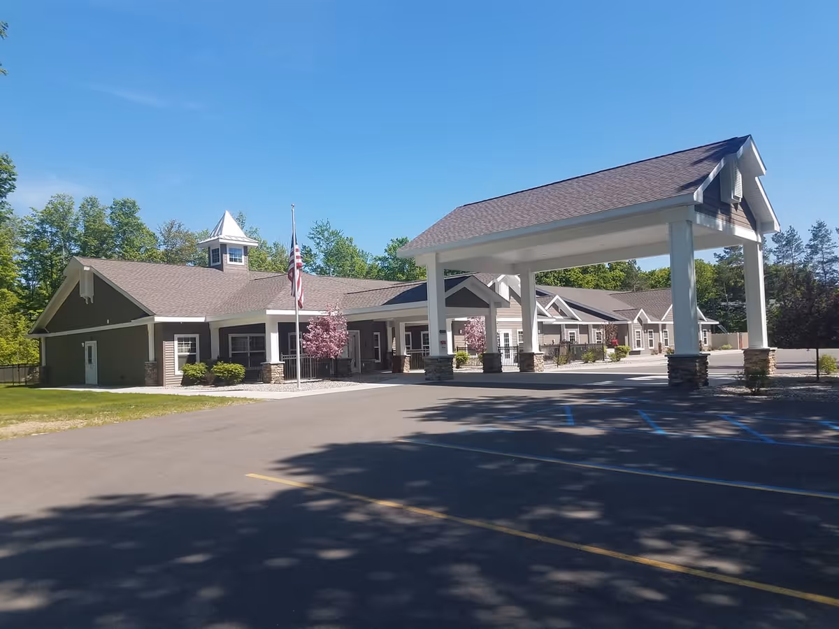 Exterior view of a single-story senior living facility building with a covered entrance, an American flag on a flagpole, and surrounding greenery under a clear blue sky.