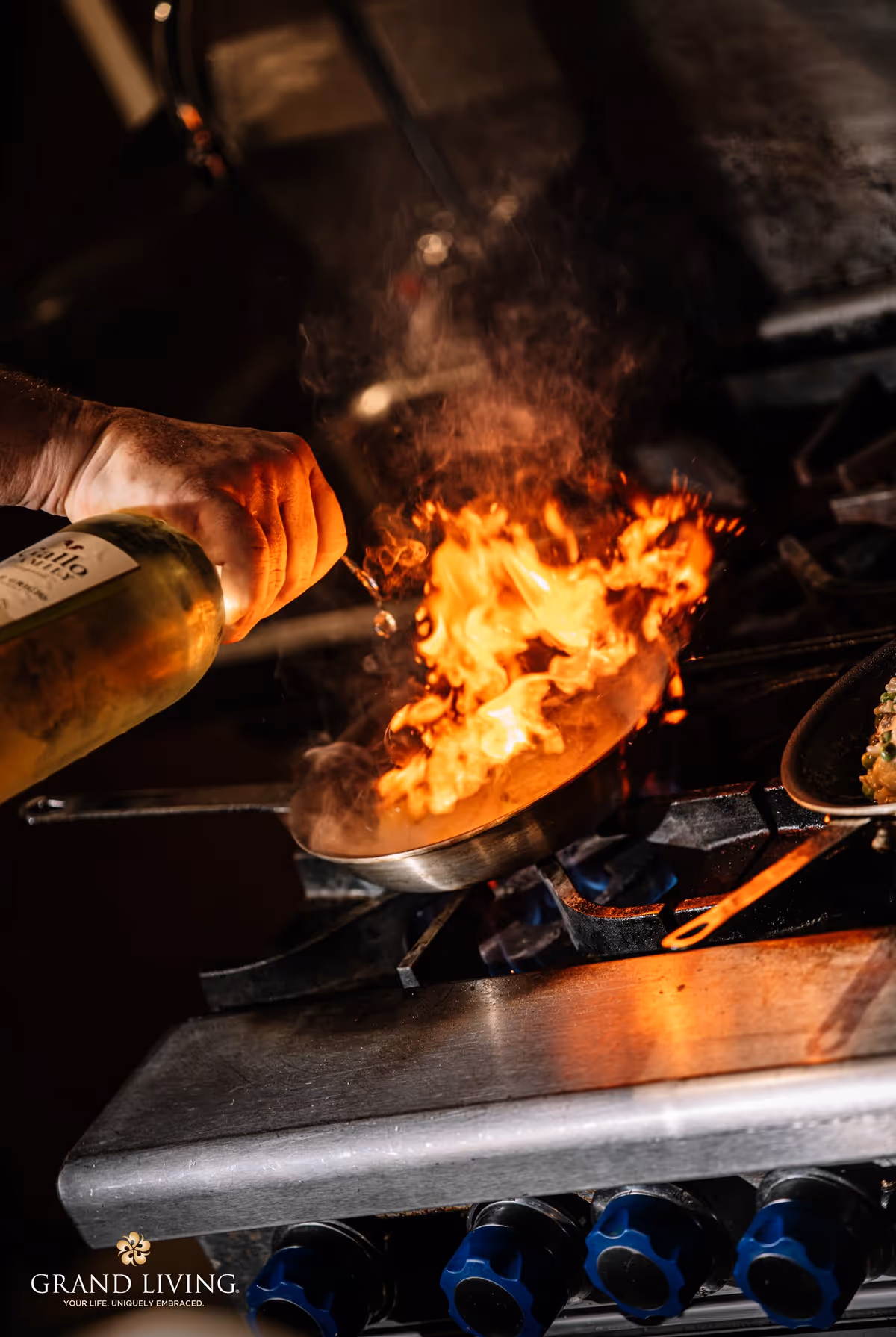 A hand pouring liquid from a bottle into a flaming pan on a gas stove in a kitchen setting, with visible flames and steam rising from the pan.