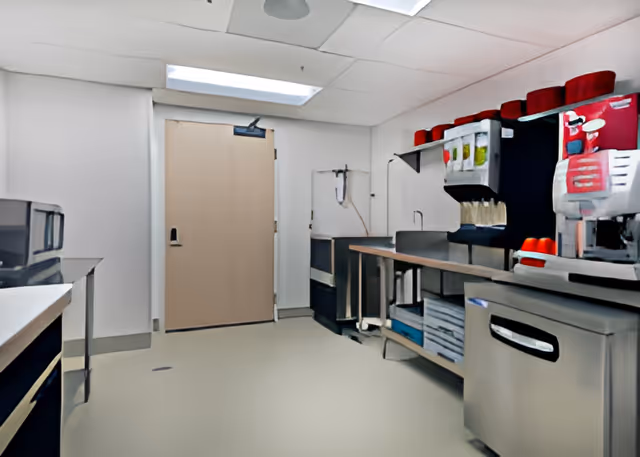 A clean, well-lit commercial kitchen area with stainless steel countertops and appliances, including a coffee machine and beverage dispensers. There is a wooden door at the back and various kitchen equipment and supplies neatly arranged.