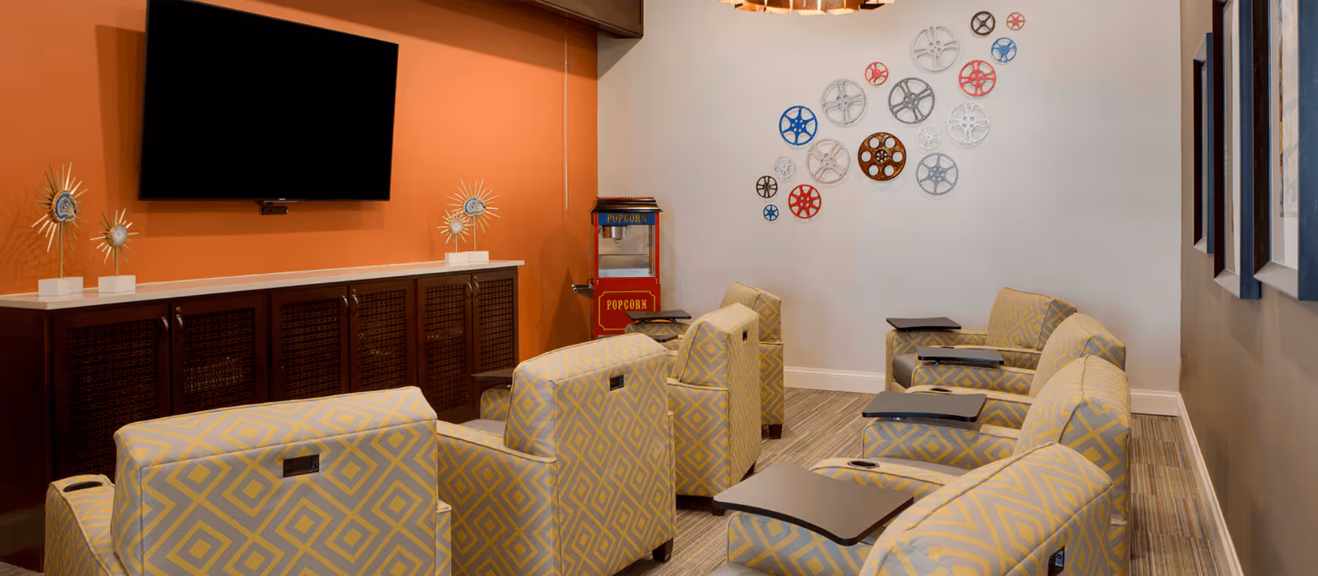 A cozy lounge area with patterned yellow and gray armchairs arranged in rows facing a wall-mounted flat screen TV. The wall behind the TV is painted orange, and there is a popcorn machine in the corner. The opposite wall is decorated with colorful film reel art pieces.