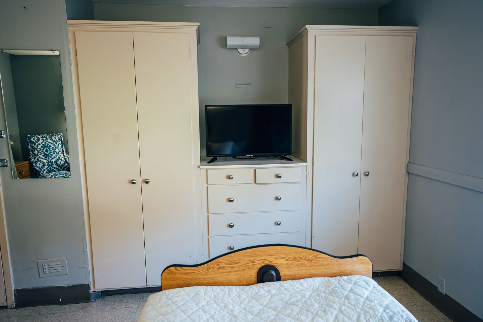 A bedroom in a senior living facility with a bed in the foreground, two white wardrobes on either side of a white chest of drawers with a flat-screen TV on top. The walls are painted light blue and there is a small mirror on the left wall.