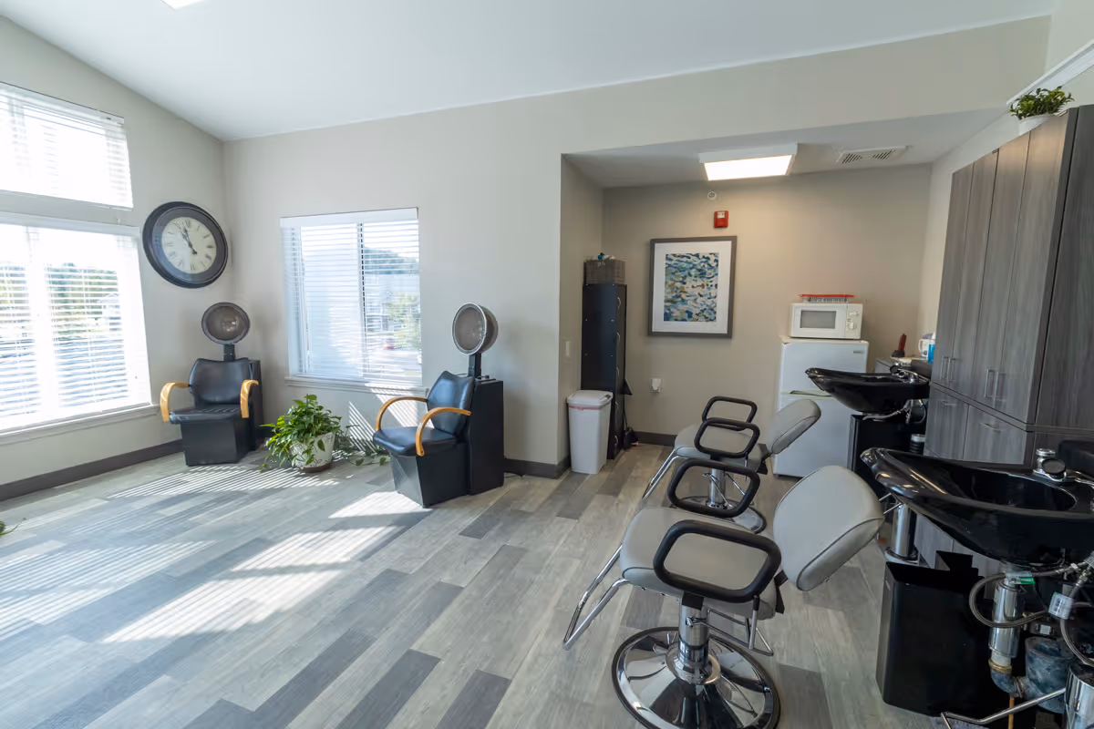 A bright hair salon area with two black hair drying chairs near large windows with blinds, two salon styling chairs facing black wash basins, a small refrigerator with a microwave on top, a trash can, a tall cabinet, and a framed artwork on the wall.