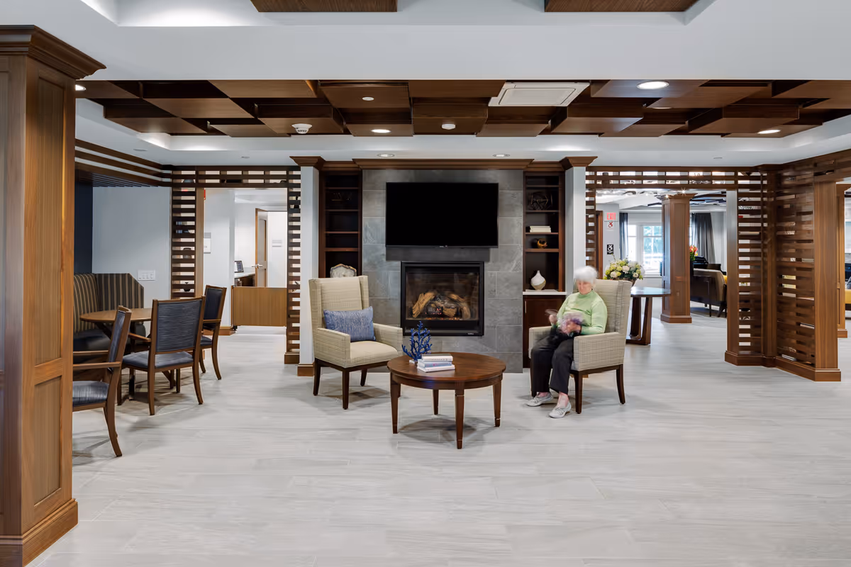 A spacious assisted living facility common area with light gray tiled flooring and wooden ceiling accents. Two beige armchairs with a small round wooden table between them are positioned in front of a modern fireplace with a mounted flat-screen TV above it. An elderly woman wearing a light green sweater and dark pants is seated in one of the armchairs. To the left, there is a dining area with a table and chairs, and the room is framed by wooden columns and slatted wood partitions.
