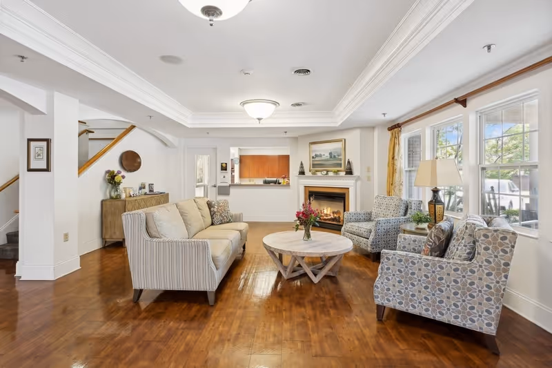 A bright and inviting living room area with wooden flooring, a beige striped sofa, two patterned armchairs, a round wooden coffee table with a vase of flowers, a fireplace with a painting above it, large windows with yellow curtains, a table lamp, and a sideboard with decorative items near a staircase.
