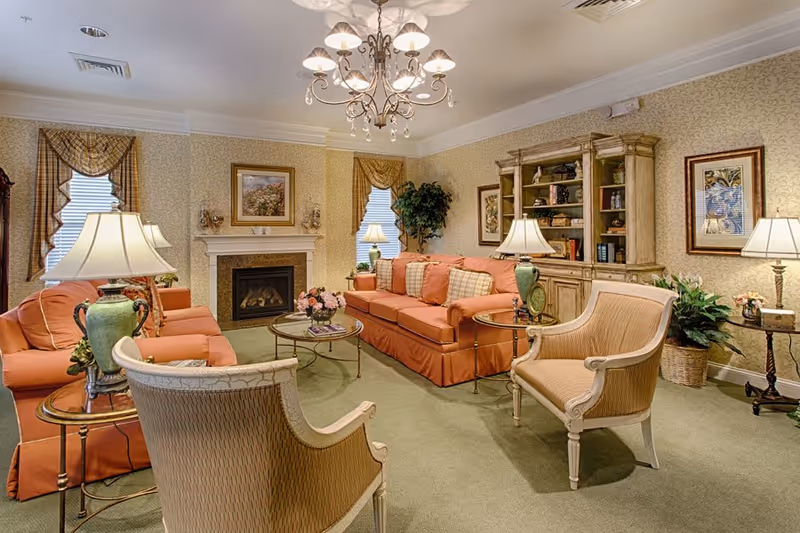 Well-appointed senior living community living room with coral sofas and armchairs arranged around a coffee table and fireplace under a chandelier.