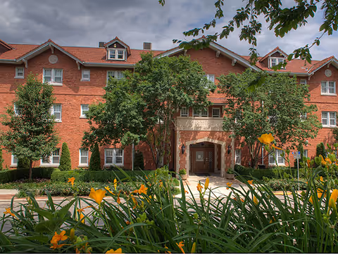 Front exterior view of Rockhill Manor, a three-story red brick building with a tiled roof, surrounded by green trees and yellow flowers in the foreground under a partly cloudy sky.