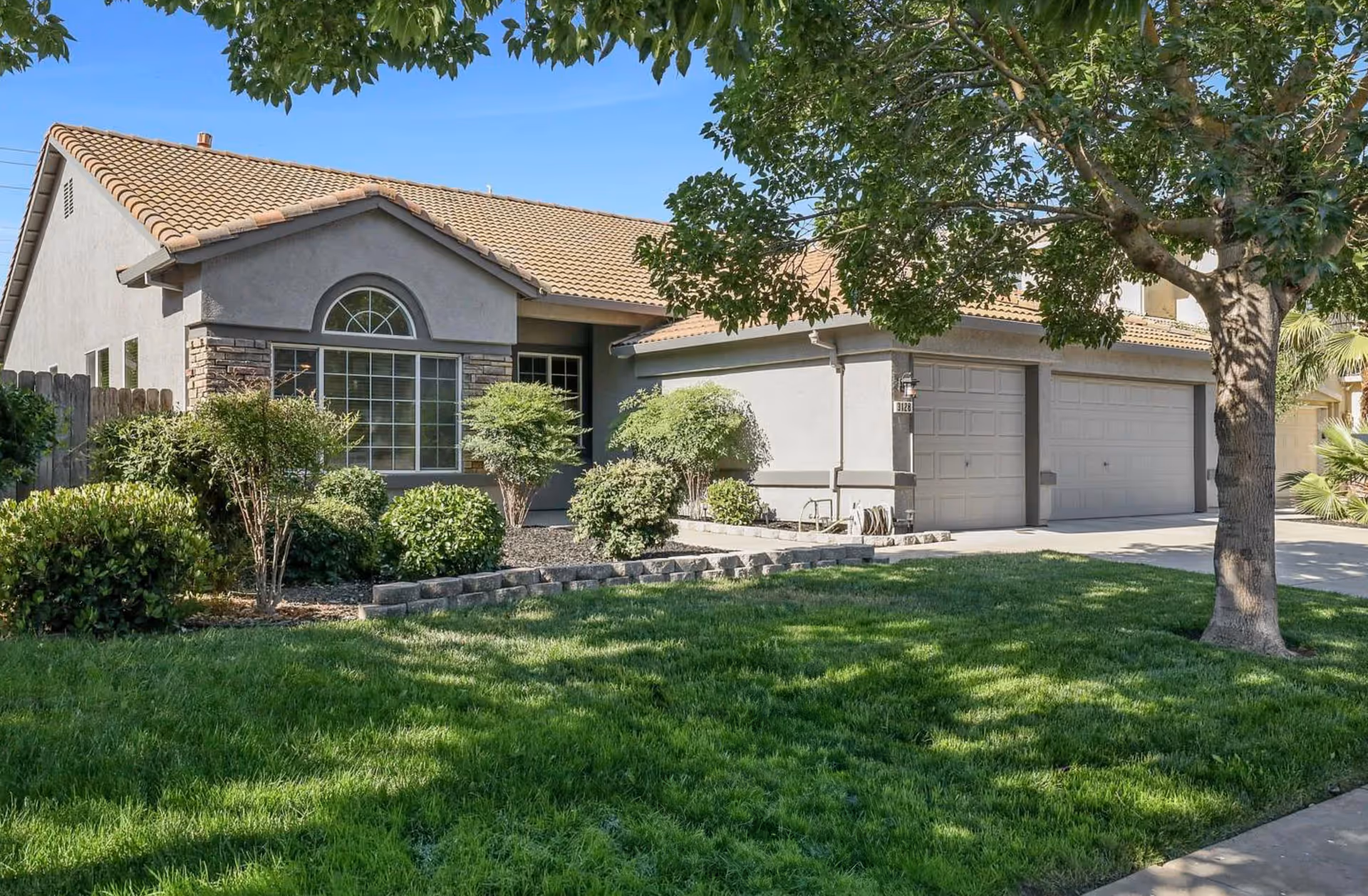 Exterior view of a single-story house with a tiled roof, a large window with an arched top, a two-car garage, and a well-maintained front yard with green grass, bushes, and a tree.