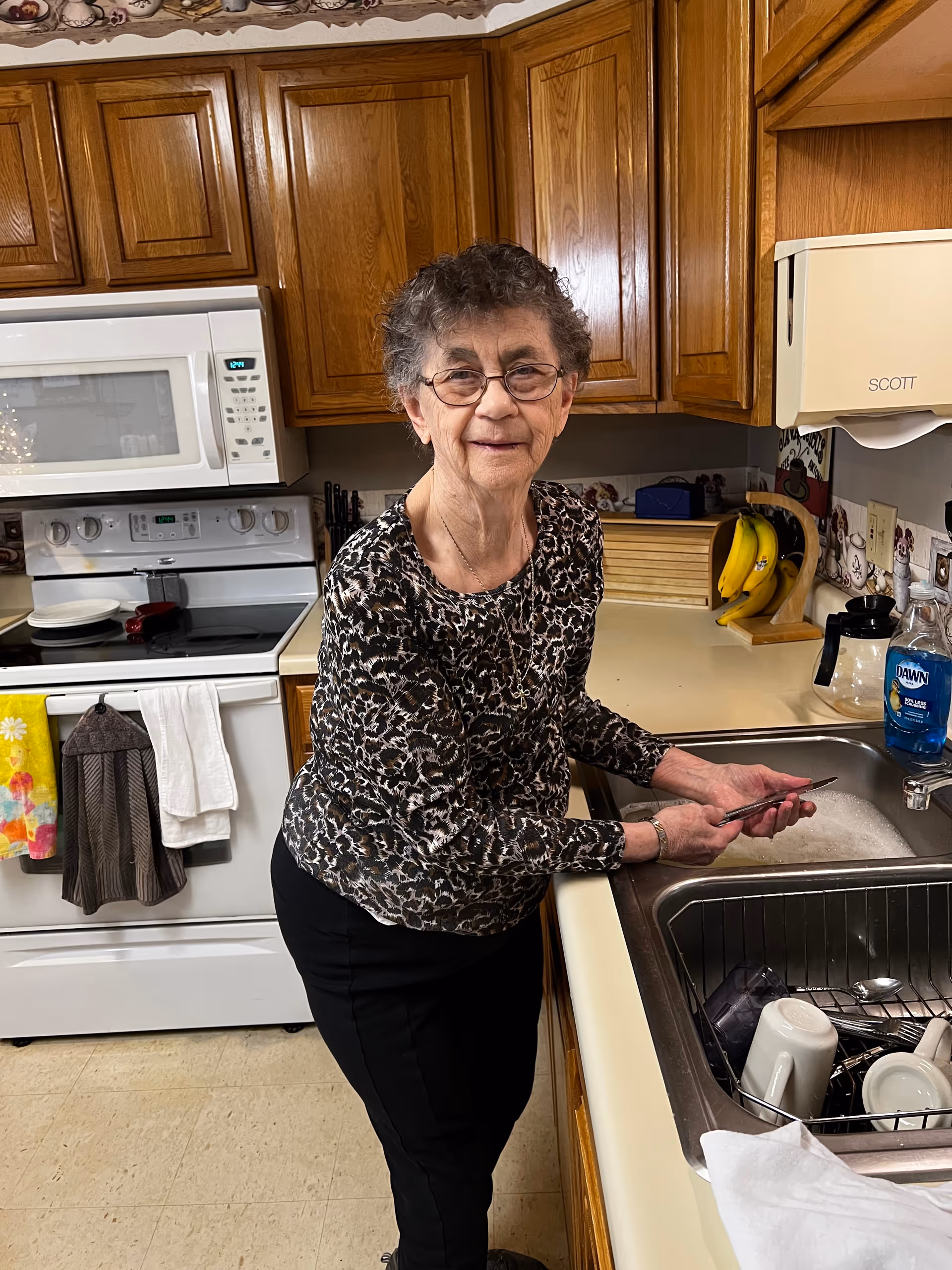 An elderly woman with glasses is standing in a kitchen washing dishes at the sink. The kitchen has wooden cabinets, a white stove with a microwave above it, and various kitchen items including bananas, dish soap, and towels hanging on the oven handle.
