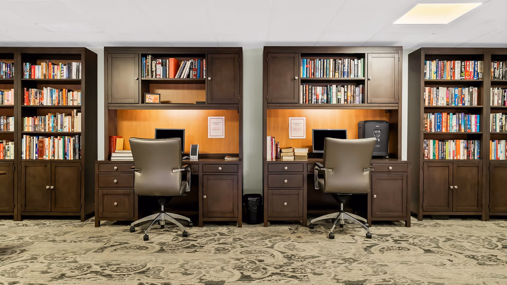 A quiet study area with two dark wood desks, each equipped with a computer monitor, office chair, and shelves filled with books. The desks are built into a wall lined with additional bookshelves filled with various books. The carpet has a patterned design, and the lighting is bright and even.