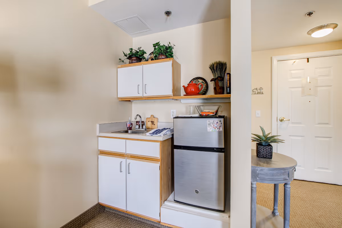 Small kitchenette area with a stainless steel mini refrigerator, white cabinets with wooden trim, a sink, and a countertop. There are decorative items on the shelf above the refrigerator, including a red teapot and plants. To the right, there is a small round table with a potted plant and a white door in the background.