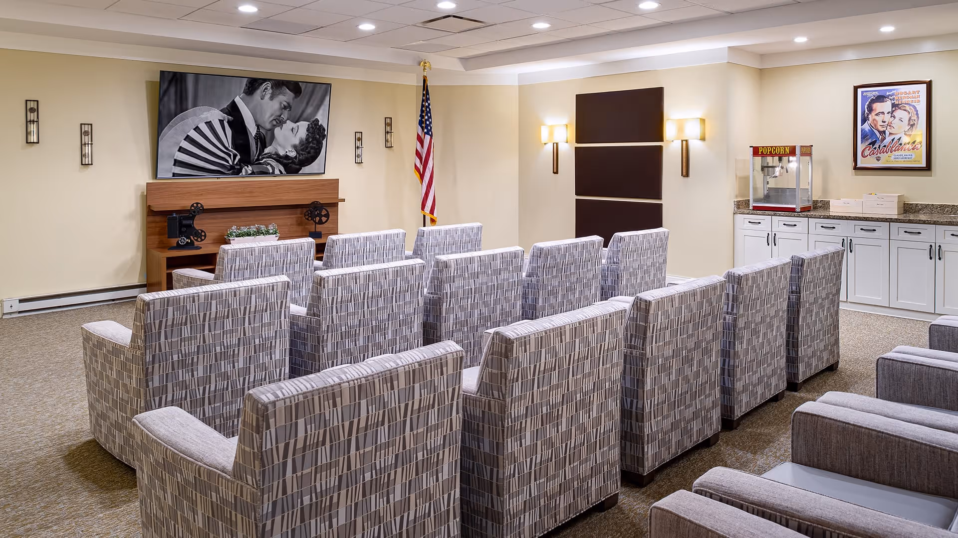 A small theater room with three rows of patterned armchairs facing a wall-mounted black and white photo of a couple kissing. There is an American flag in the corner, a popcorn machine on a countertop, and a framed Casablanca movie poster on the wall.