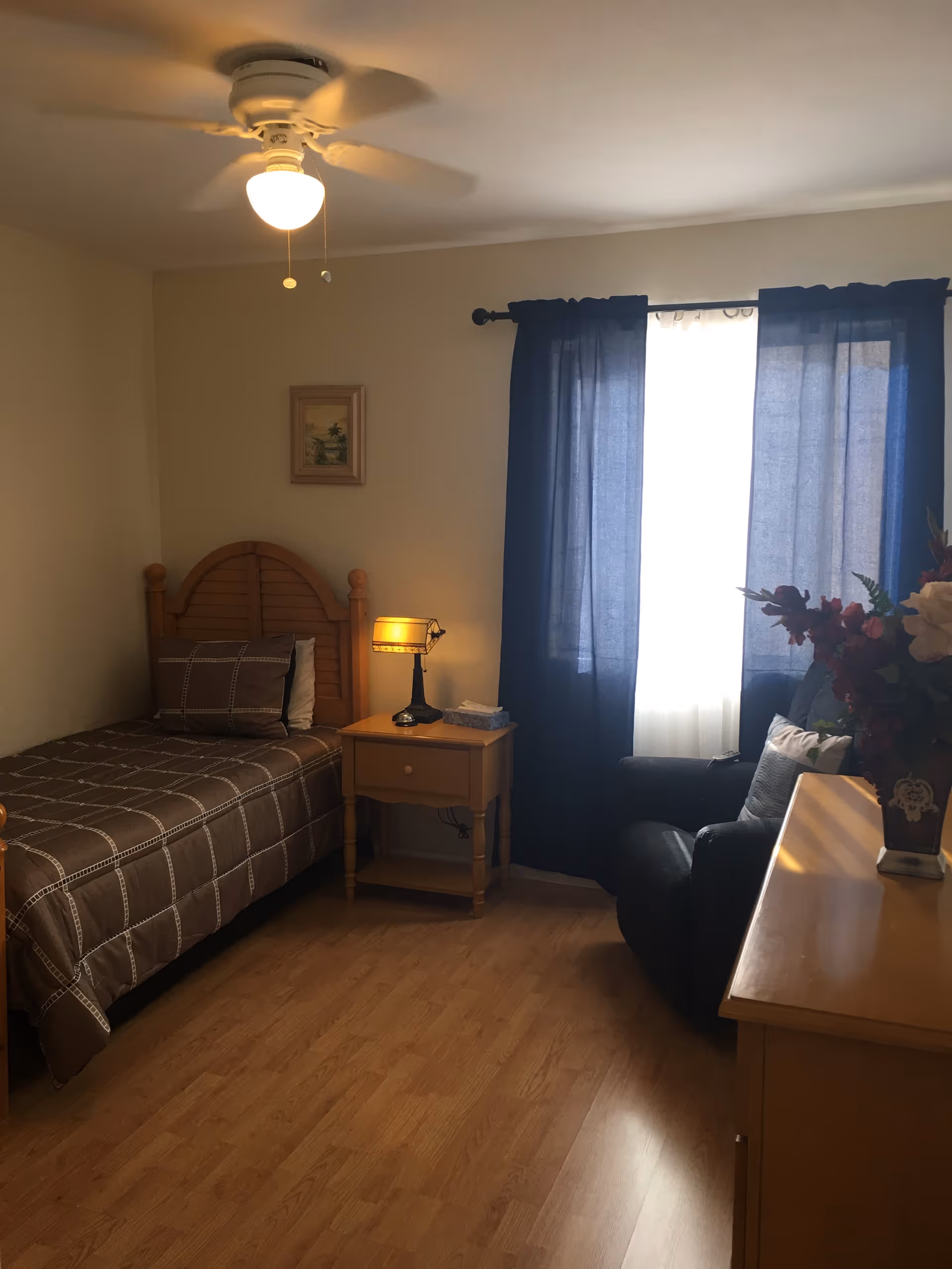 A small bedroom with a single bed covered in a brown checkered bedspread, a wooden nightstand with a lamp and tissue box, a window with blue curtains, a black armchair with pillows, a wooden dresser with a vase of flowers, and a ceiling fan with a light.
