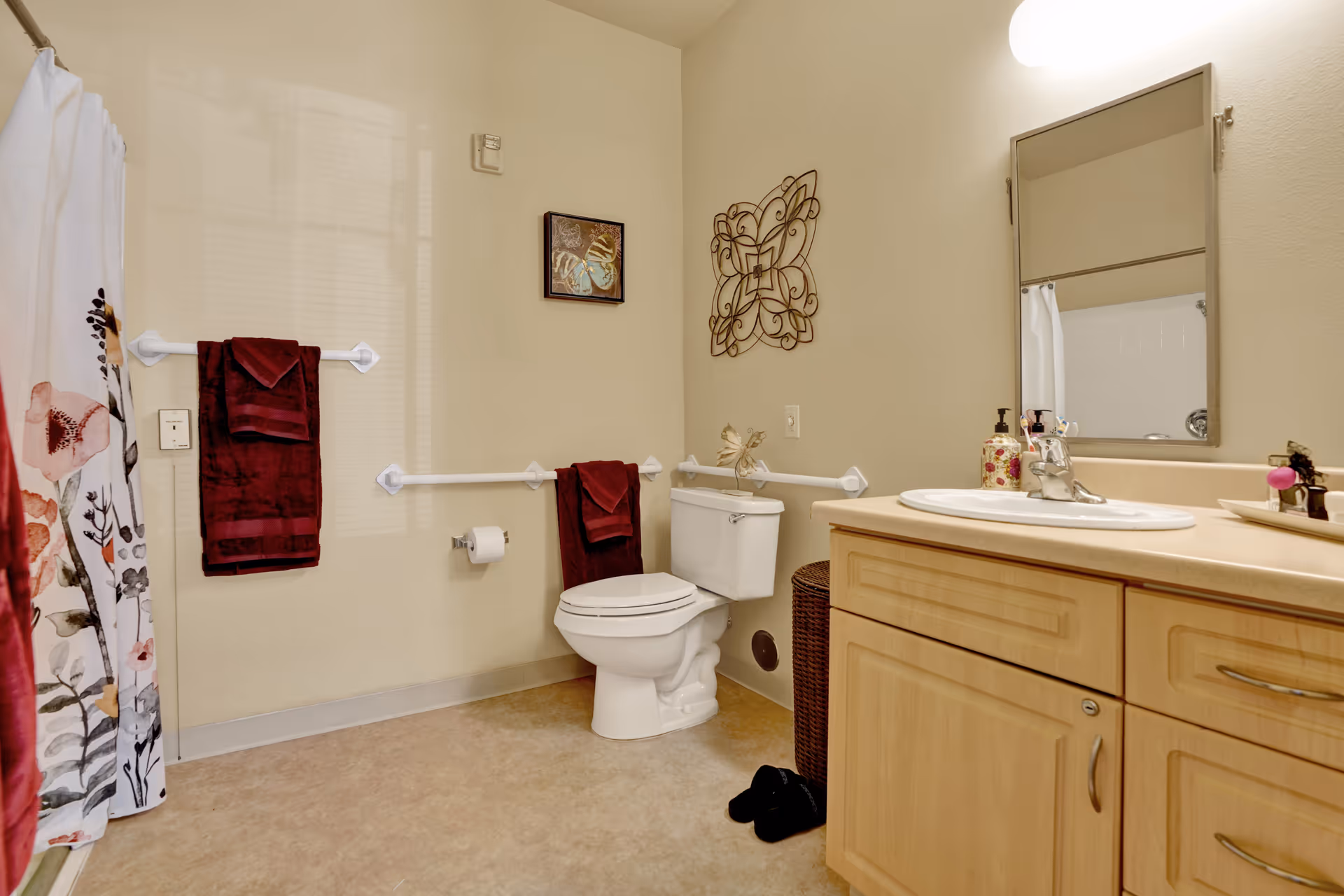 A bathroom with beige walls and floor, featuring a toilet with grab bars on both sides, a wooden vanity with a sink and mirror above it, a floral shower curtain, and burgundy towels hanging on the wall. There is a decorative wall art above the toilet and a wicker laundry basket beside it.