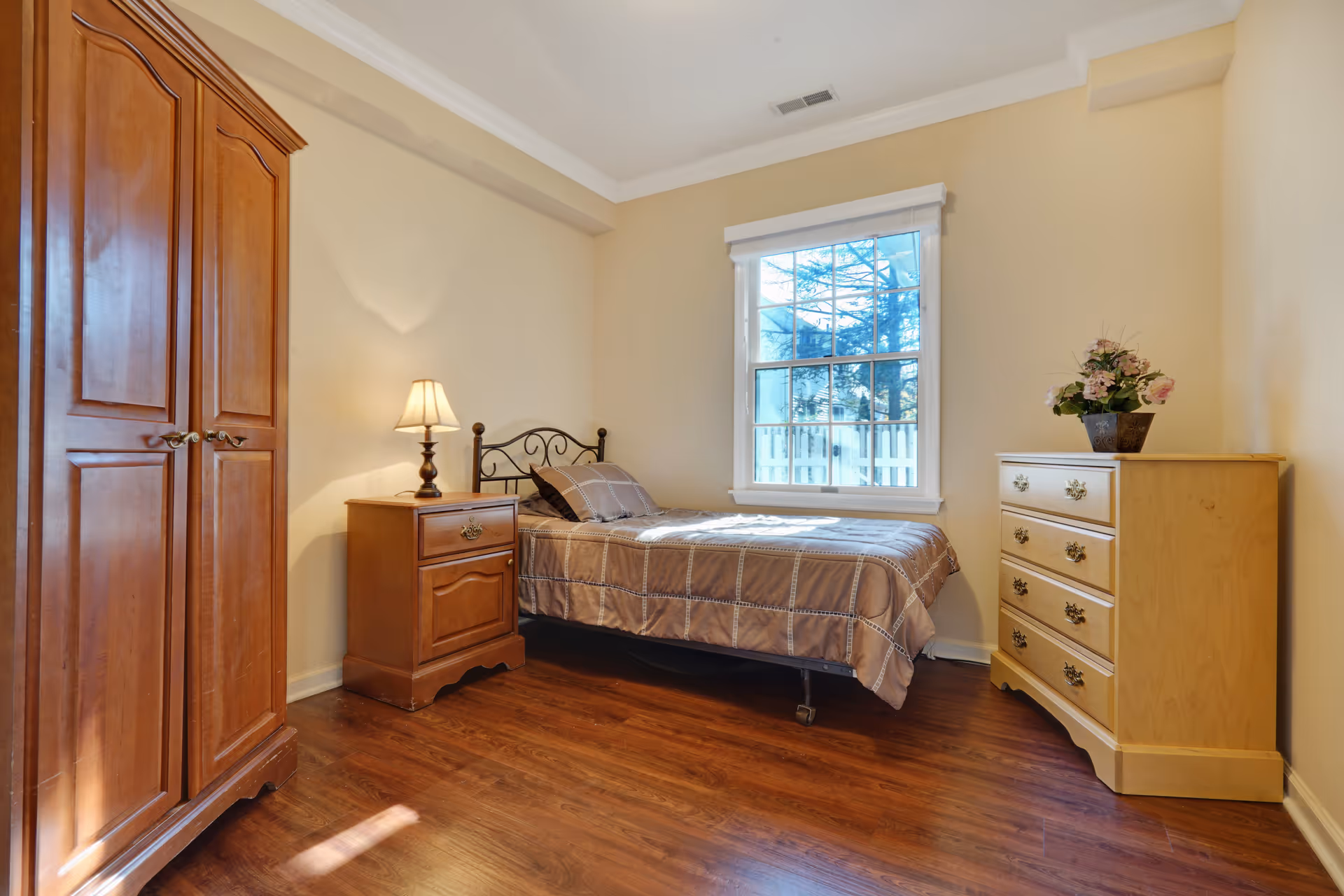 A small bedroom with a single bed covered in a brown and beige checkered bedspread. Next to the bed is a wooden nightstand with a lamp on it. There is a wooden wardrobe on the left side of the room and a light-colored wooden dresser with a flower pot on top on the right side. A window with white trim lets in natural light, illuminating the wooden floor and beige walls.