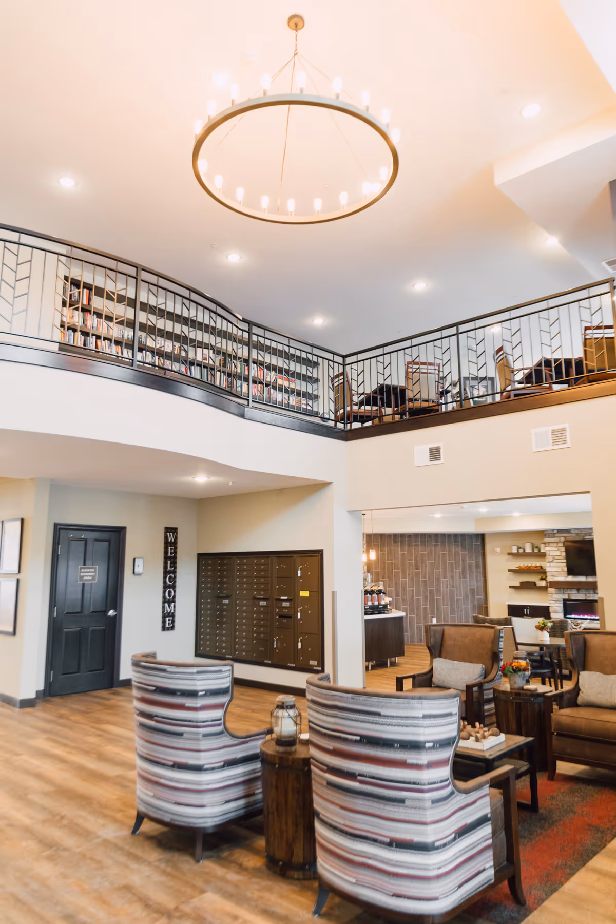 Interior view of a senior living facility lobby area with a high ceiling and a large circular chandelier. There are two striped armchairs facing a small wooden table with a lantern on it. In the background, there is a mail area with multiple mailboxes and a black door with a 'Welcome' sign on the wall. Upstairs, there is a balcony with a railing and bookshelves, along with some chairs and tables. The space has warm lighting and wooden flooring.