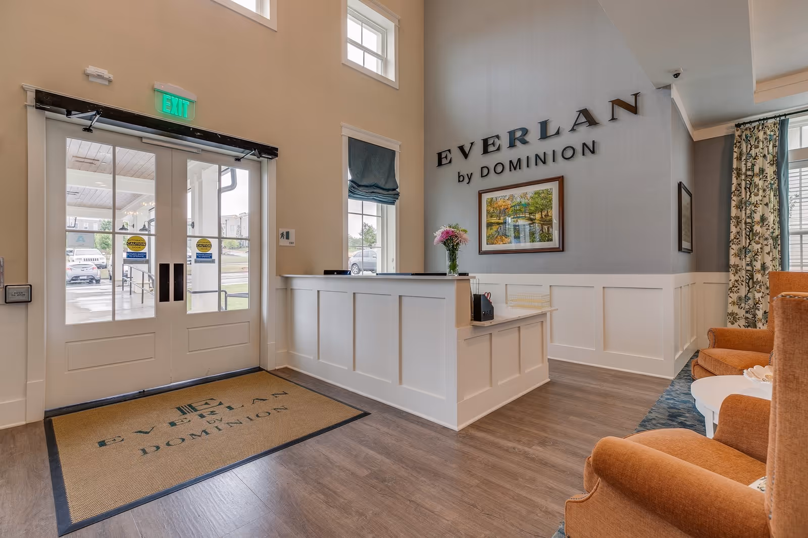 Reception area of Everlan by Dominion featuring a white reception desk with a vase of flowers, a framed painting on the wall, large windows with blue roman shades, and a seating area with orange armchairs and a small white table. Double glass doors with exit signs lead outside.