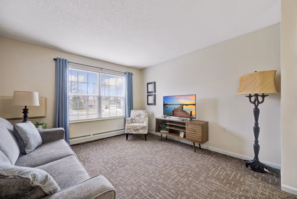 A bright living room with a large window covered by blue curtains, a gray sofa with pillows, a patterned armchair, a wooden TV stand with a flat-screen TV displaying a sunset over a dock, and two lamps, one table lamp and one floor lamp.