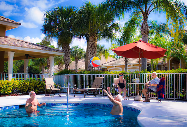 Two elderly men playing with a beach ball in a swimming pool while two elderly people sit on chairs under a red umbrella near the poolside, surrounded by palm trees and a sunny sky.