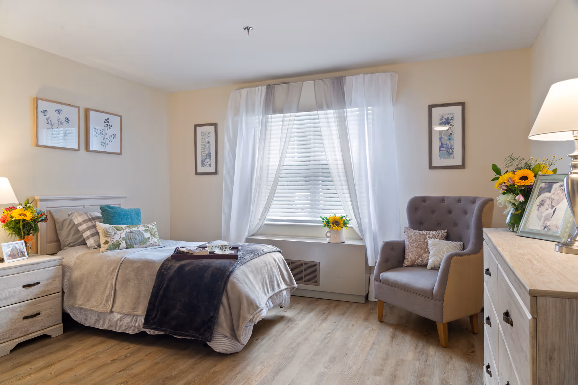 A cozy bedroom with a single bed made with beige and gray bedding, a dark throw blanket, and decorative pillows. There is a wooden nightstand with a lamp, flowers, and a framed photo. A window with white sheer curtains lets in natural light. A gray upholstered armchair with two pillows sits next to a wooden dresser with a lamp, flowers, and a framed picture. The walls are decorated with framed botanical prints.
