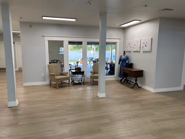 A spacious interior room with light wood flooring and white walls. Two patterned armchairs are positioned near a large window that looks out onto an outdoor patio with tables and chairs. A small wooden table with a mannequin dressed in a blue outfit and three floral paintings on the wall are also visible. The room is well-lit with ceiling lights.