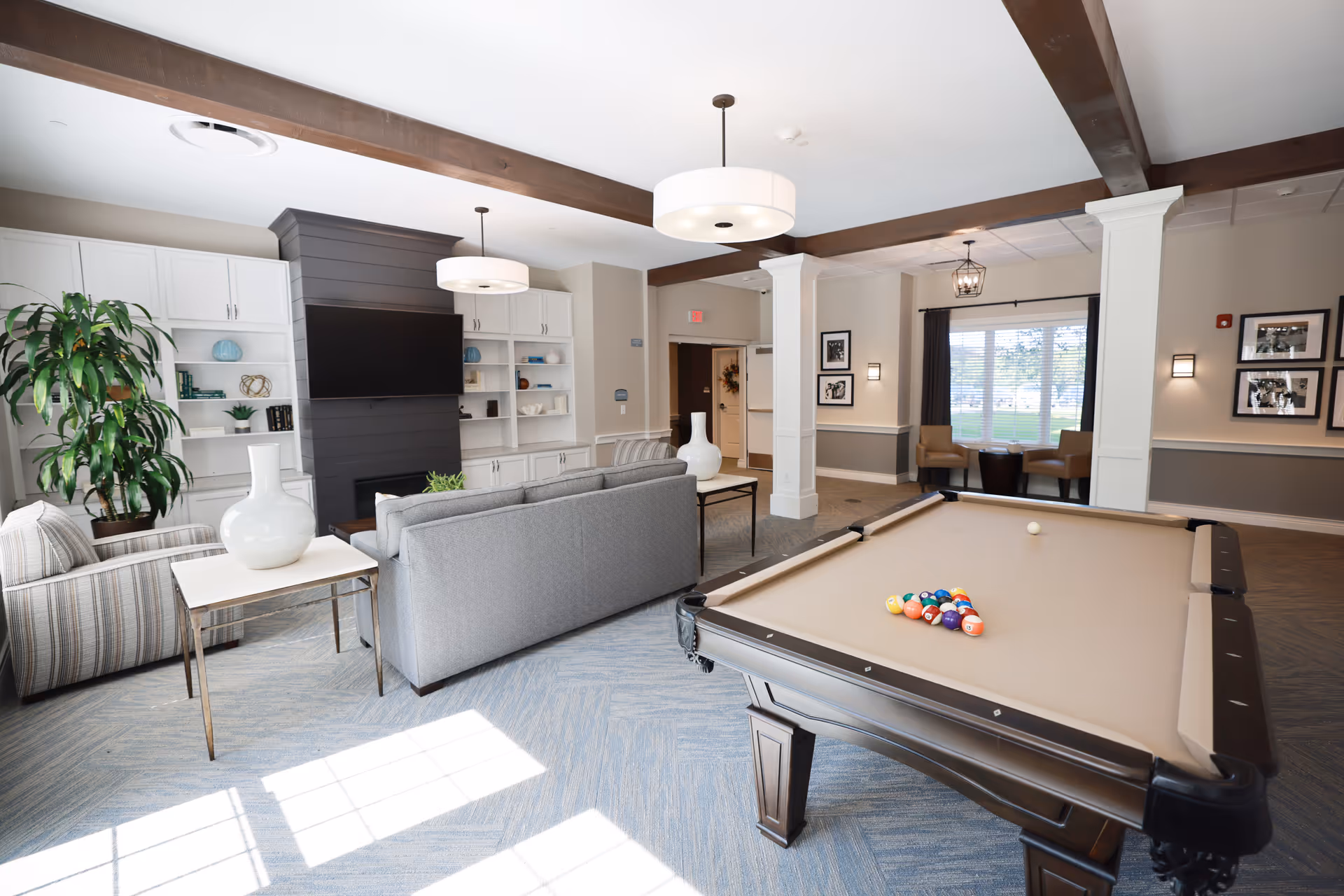 Communal living room featuring a pool table in the foreground, gray sofas, built-in shelves with a TV, and large windows.