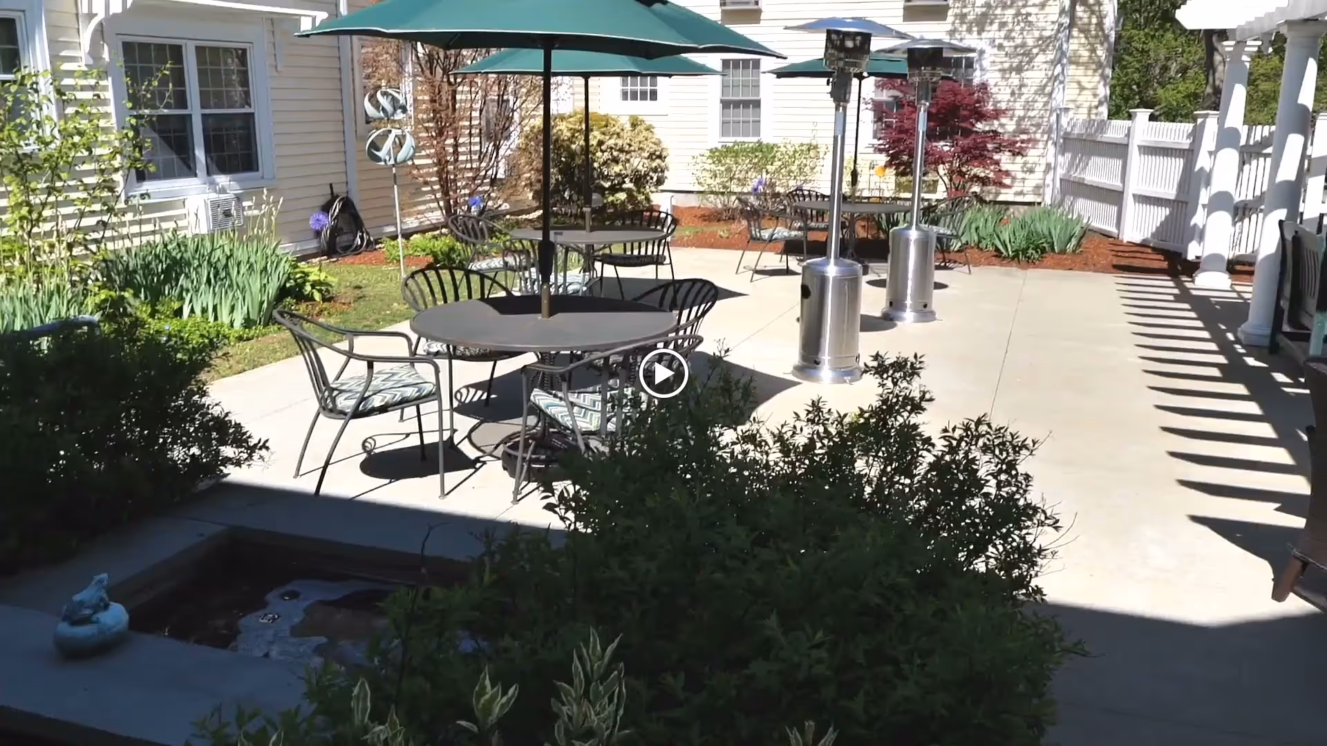Sunlit outdoor patio courtyard with round tables, green umbrellas, patio heaters, chairs, and surrounding landscaping.