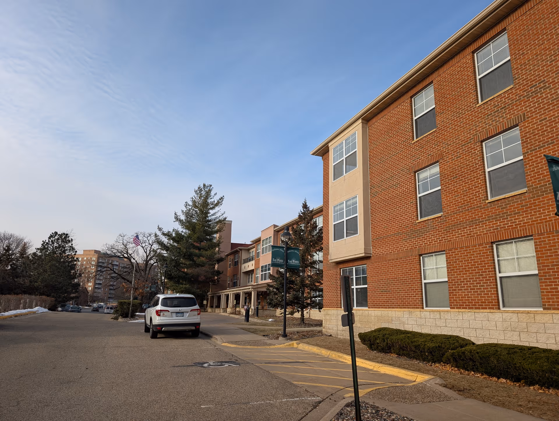 Exterior view of a senior living facility named The Pines Senior and Assisted Living. The building is made of red brick with multiple windows. A white SUV is parked on the driveway in front of the building. There are trees and bushes along the sidewalk, and a clear blue sky above.