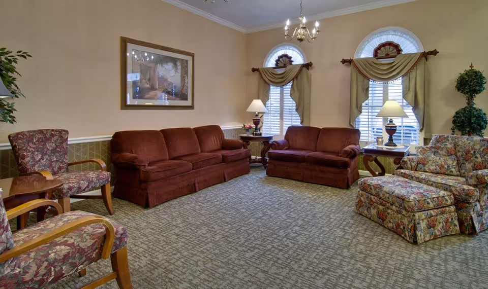 A cozy senior living facility common area with two maroon sofas, two floral armchairs, a floral ottoman, two side tables with lamps, and two arched windows with beige curtains and white shutters. The walls are beige with a framed picture hanging above one sofa and a potted plant in the corner.
