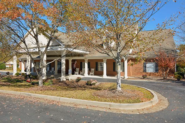 Front entrance of a brick senior living building with a covered porch, white columns, rocking chairs and trees in the foreground.