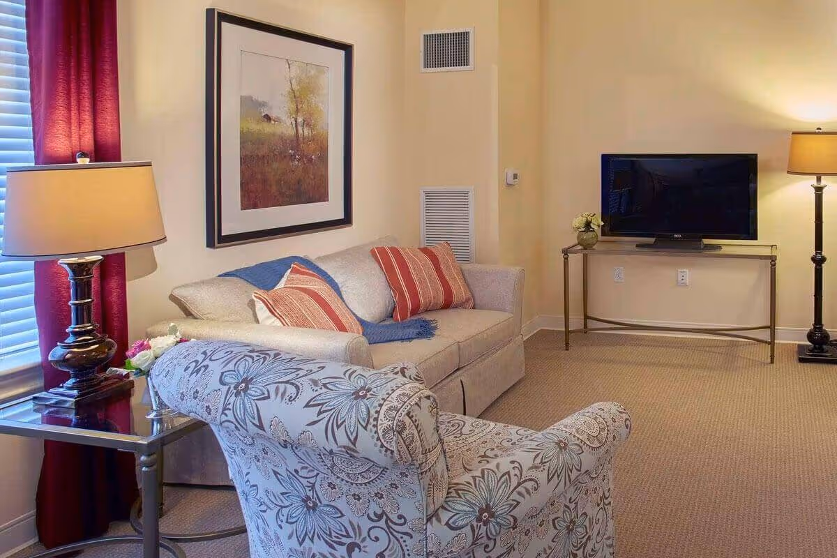 Cozy living room with a sofa, patterned armchair, table lamps, framed art, and a TV on a console.