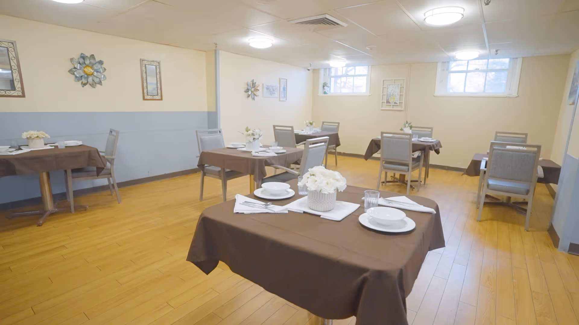 Small dining room with multiple tables covered in brown tablecloths set with white dishes and floral centerpieces.